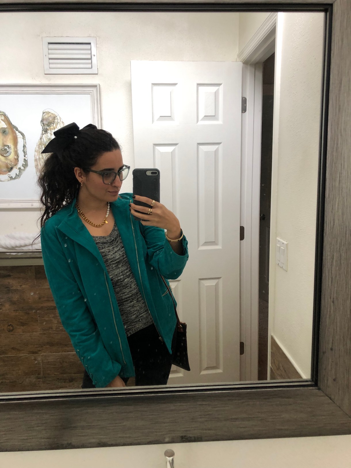Kayla Kumari Upadhyaya wears a black and white knit blend sweater and black jeans and a teal suede motojacket. Her hair is in a ponytail with a black bow and she wears blue framed glasses and a chunky gold chain and pearl necklace.