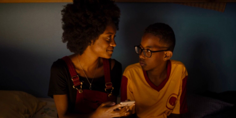 A young woman sits on a bed next to her little brother in orange lighting.