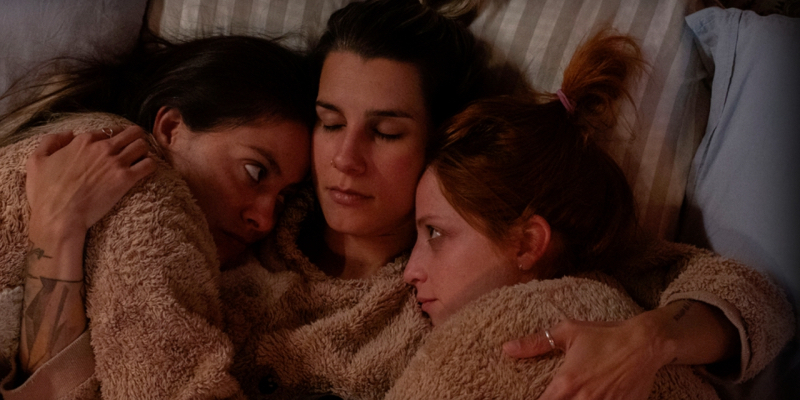 A woman lies in bed with her arms around her two girlfriends who are making eye contact.