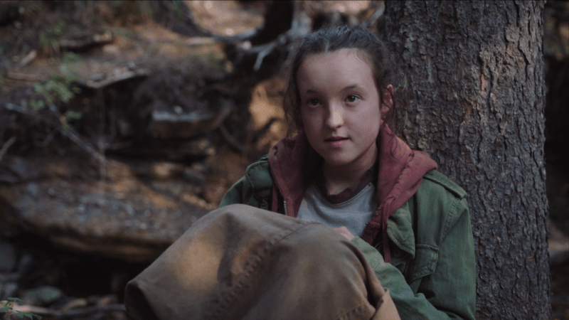 The Last of Us: Ellie sits against a tree in the forest with Joel's jacket on her lap, looking sassily up at her travel companion.