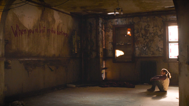 The Last of Us: Ellie sits alone, small in a big room, with the words "WHEN YOU'RE LOST IN THE DARKNESS..." scrawled across the wall in red spray paint.