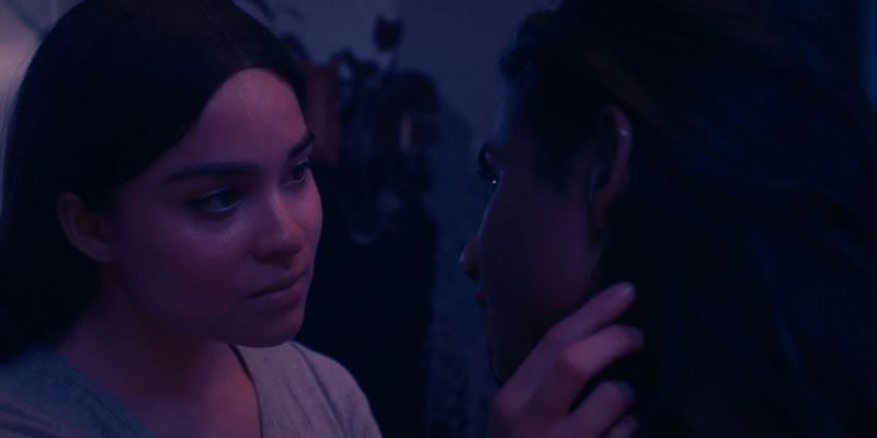 Devery Jacobs as Kawenniióhstha looks at Priya Guns as Malai in soft pink and blue lighting with one hand touching her hair.