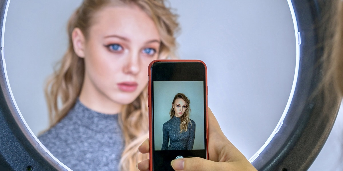 A blonde woman with blue eyes is filming a TikTok while making a sad pouty face. She is framed by a ring light, against a blue wall.