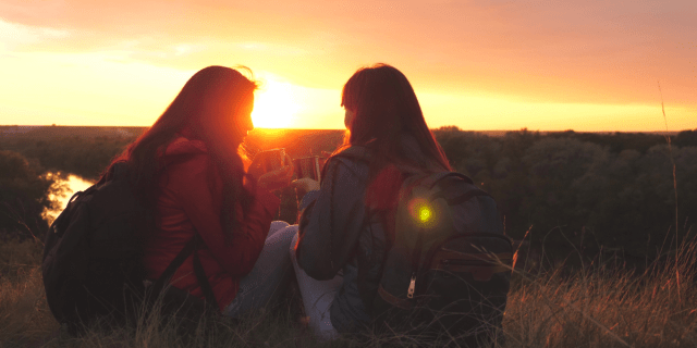 The silhouettes of two women are visible on a hill during sunset. They both have long, dark hair, wear backpacks and jeans, and hold metal cups in their hands. They sit on the ground and face each other. The woman on the left wears a red jacket and the woman on the right wears a blue jacket.