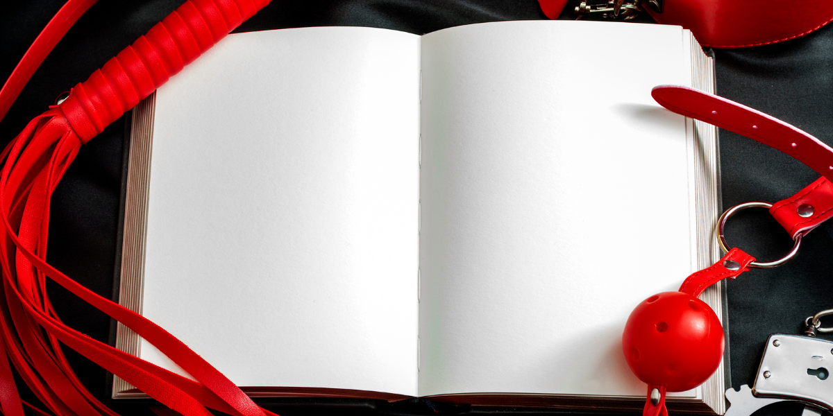 A blank, unlined journal is open on a black sheet. A red flogger is on the left of the journal and a red ball gag is on the right of the journal.