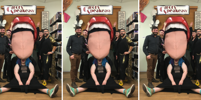 This image features a giant 3D tongue art installation that used to hang at Carol's Speakeasy. The lips are red and shiny, the teeth are large and white, and the tongue is pink and sticking out of the mouth. Five queer volunteers at the archive pose around the tongue, smiling and laughing with this piece of queer history.
