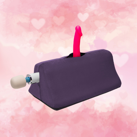 Against a white and pink heart-covered background, there is a purple wedge pillow with a pink dildo mounted in the top pocked and a Magic Wand vibrator mounted in the pocket on the left side.