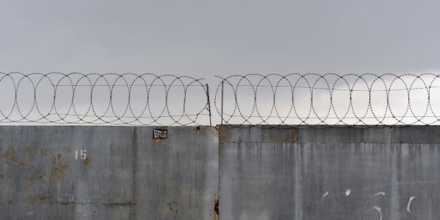 The top of a concrete wall is visible against a gray sky. On top of the wall, there is looped barbed wire along with barbed wire that's running horizontally. On the left side of the wall, the number 15 is stenciled in white paint.