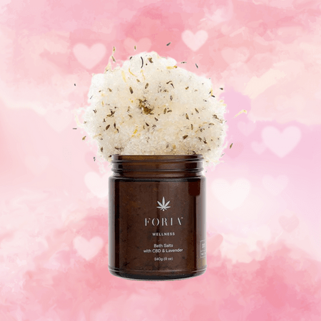 Against a white and pink background covered in hearts, there is a brown glass jar with an image of a white marijuana leaf. Text on the jar reads, "Foria, Wellness Bath Salts with CBD and Lavender." The white salts with small dark flecks are exploding out from the top of the jar.