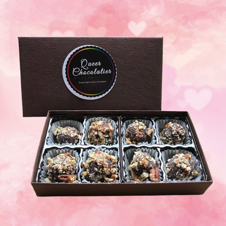 Against a white and pink background covered in hearts, there is a brown, open box containing eight, nut-covered chocolate truffles. The lid of the box is propped up in the background. Text on the box reads, "Queer Chocolatier."