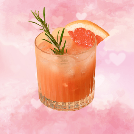 Against a white and pink background covered in hearts, there is a short glass containing a pink beverage. It's garnished with rosemary and a slice of grapefruit.