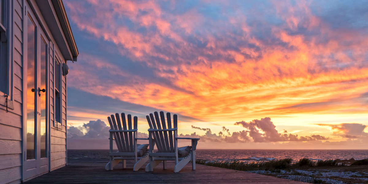 Two white wooden chairs sit on a deck that's attached to a white house on the left. The chairs face pink and orange sunset and a large body of water.