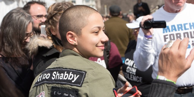 X Gonzalez is smiling to the side of camera while outside in a green jacket and shaved head, at a March For Our Lives protest.