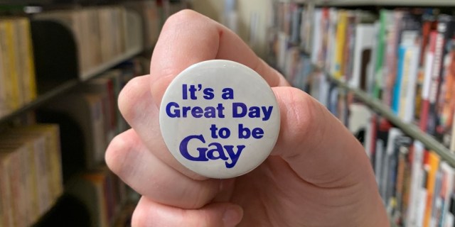 a white hand holds a button that reads "It's A Great Day To Be Gay," and we can tell they're in the archives, with many books and bookshelves behind them