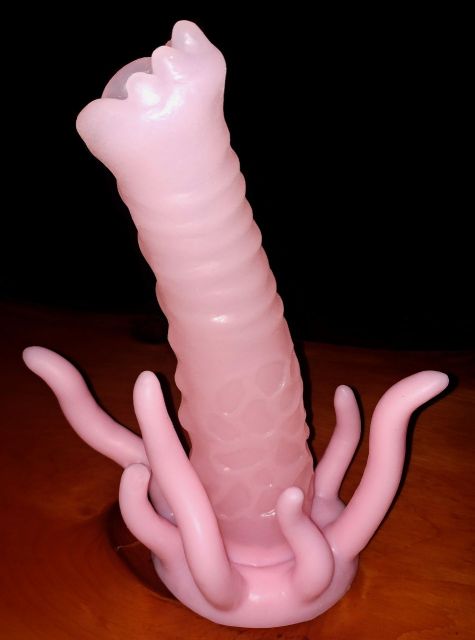 The G'lorp, a long, hollow pink dildo with ridges around the shaft and tentacles extending off of the base, sits on a red table in a dark room.