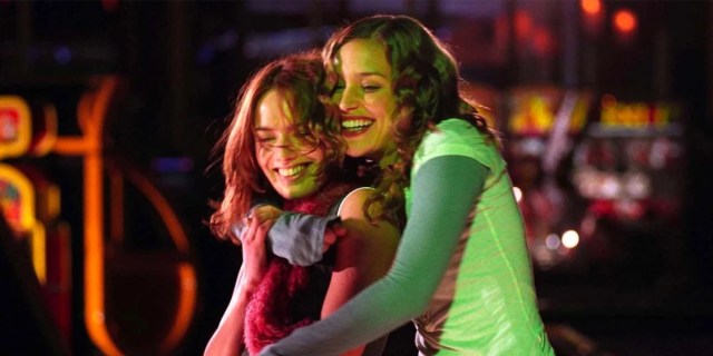 In a still from the movie Imagine Me & You, two white lesbians hold each other in a hug while laughing. They are wearing classic 00s fashion, including a useless scarf and a short sleeved t-shirt over a long sleeved t-shirt.