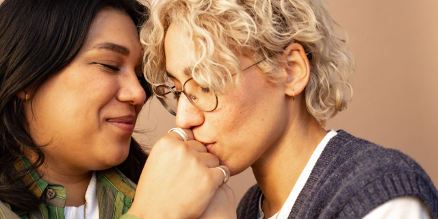 A couple, one with long dark hair and eyeliner, and another with glasses and short blonde hair who is kissing the knuckles of the other person.
