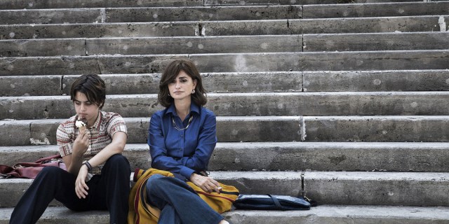 Luana Giuliani and Penélope Cruz in 1970s clothes sit next to each other on grey steps.