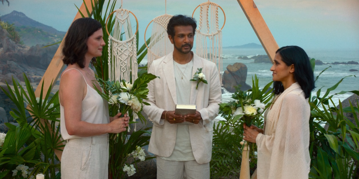 A scene from The Drop. Two women stand at an altar in white with a man between them also in white. They are under an arch on the beach, getting married by the man.