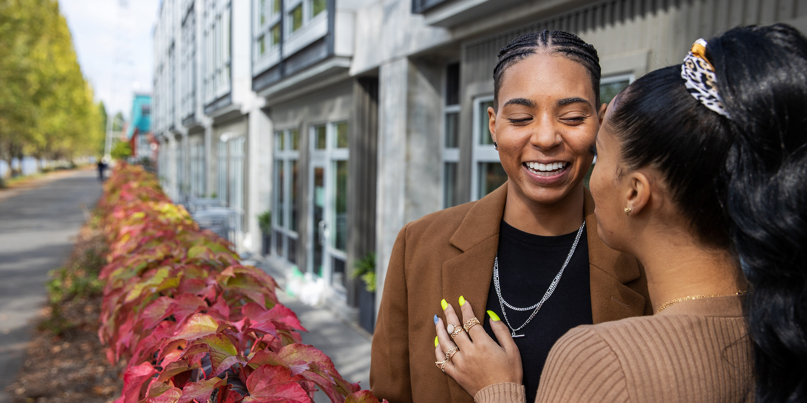 two Black queer people flirting with each other outside; one faces away from us and all we can see if her high ponytail and her neon yellow manicure touching the other person's chest; they have cornrows, a black shirt, a brown blazer, a silver chain, and a huge smile on their face