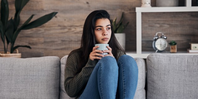 a sad woman sitting on a sofa with a mug of tea looking depressed and alone