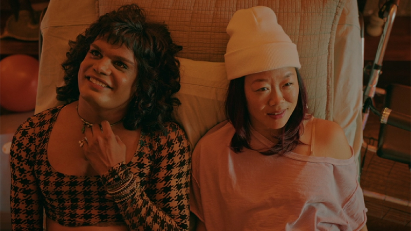 Sabi and Bessy lie side by side on Bessy's hospital bed under orange-toned lights. Sabi's wearing a tooth and fang crop top and toying with their necklace. Bessy dons a beanie and a loose, off-the-shoulder pink top. They're looking away from each other and smiling, as they reflect on relationships.