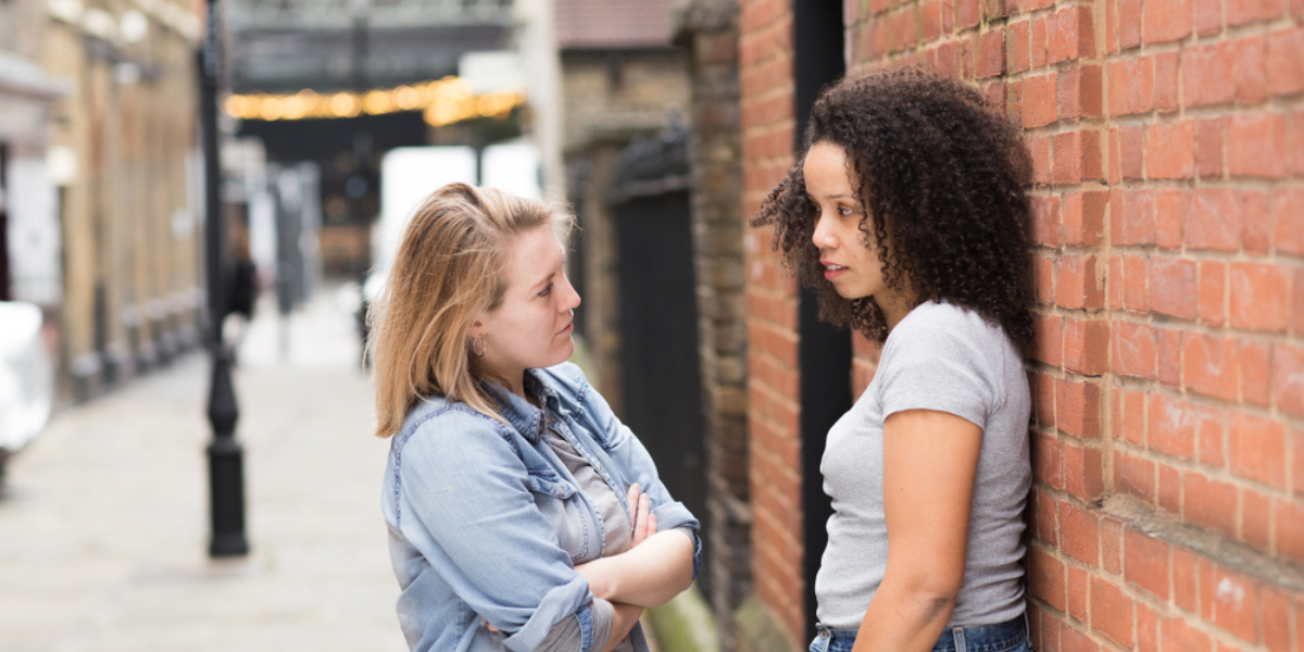 two women looking at one another not too happily. one is leaning against a brick wall.