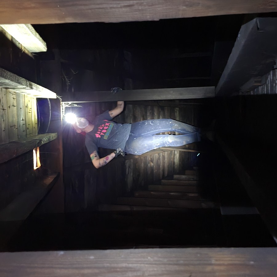 Sadie balances on a joist in the attic while wearing a headlamp. The attic is dark and wooden.