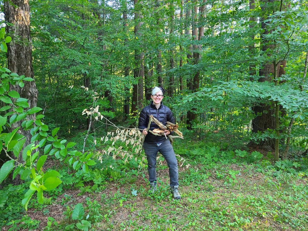 Nico stands in a jacket and jeans and hiking boots at the edge of a forest, holding a bundle of sticks and smiling.