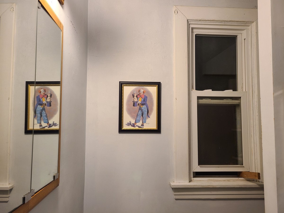 a print of a clown pulling a rabbit out of a hat is set in a stark, bleak bathroom with peeling paint. there is a window being held open with a board, blackness beyond it. the clown is ominously reflected in the mirror.