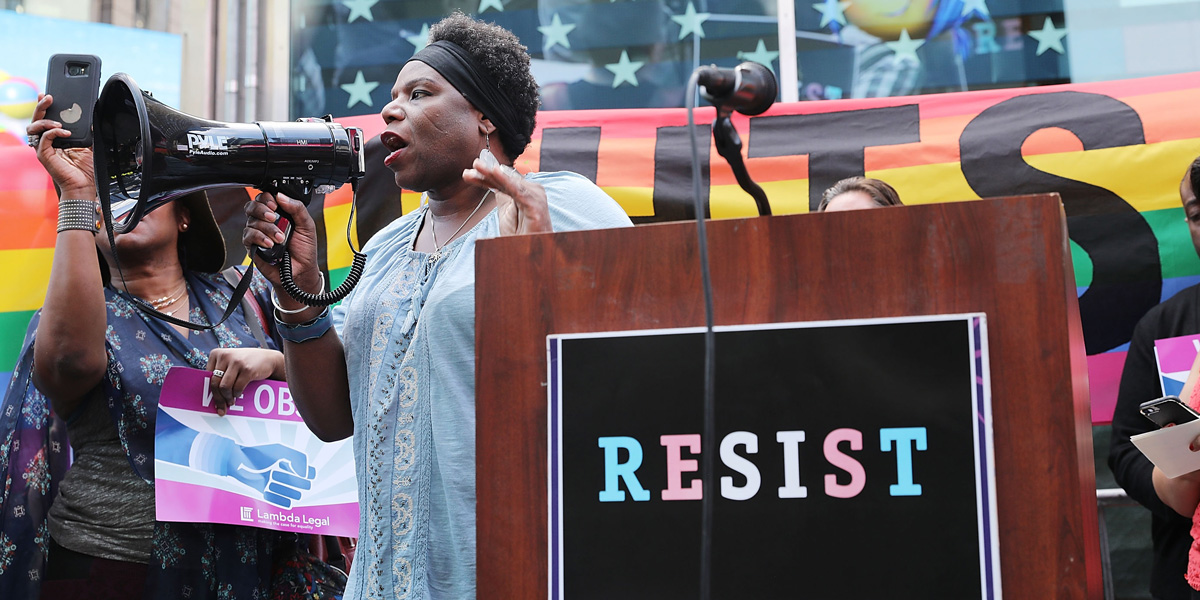 A black trans woman is at a microphone at a busy protest with a pride flag hanging behind a podium that reads "resist" in pink and blue letters (the trans pride colors)