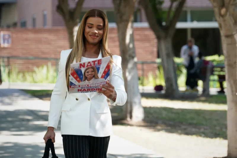 All America Homecoming: As she walks across campus, Nate looks down proudly at his campaign flyers for SGA president. She's wearing a white blazer, black pin-stripe pants, and carrying a black bag in her right hand. Her flyers say "Nate for SGA president. Working Hardin for You."