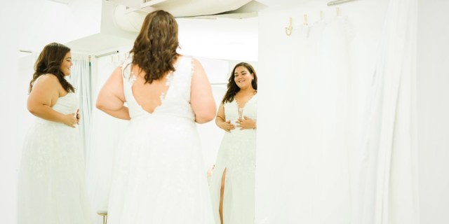 A fat bride with brown hair curled at the edges around her shoulders is in a white gown and looking at herself in the mirror of a dressing room. She is smiling and happy.