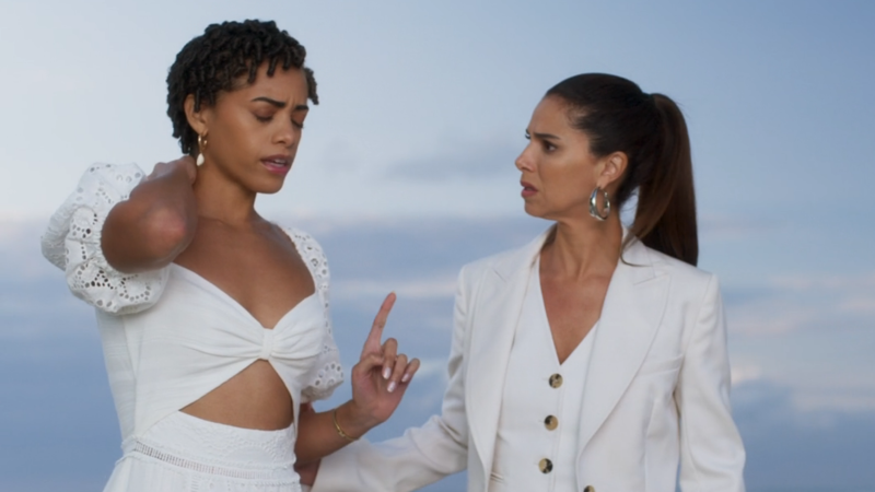 Ruby and Elena, both dressed all in white, stand on the backdrop of a beautiful ocean. Ruby closes her eyes and puts one finger up as if to as Elena to wait, and the other hand is on her tattoo. Elena looks concernedly at Ruby.