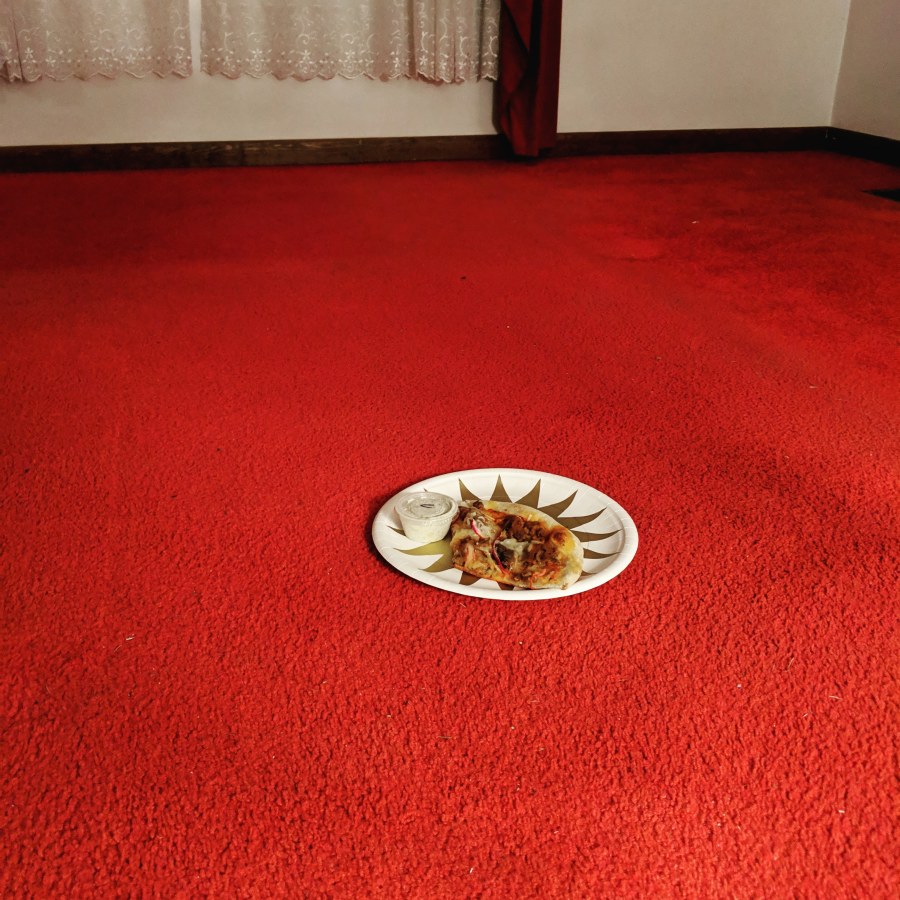 a starkly lit image of a red carpet with a piece of vegan pizza on a paper plate on it, in the center. there is a glimpse of lace curtain in the background.