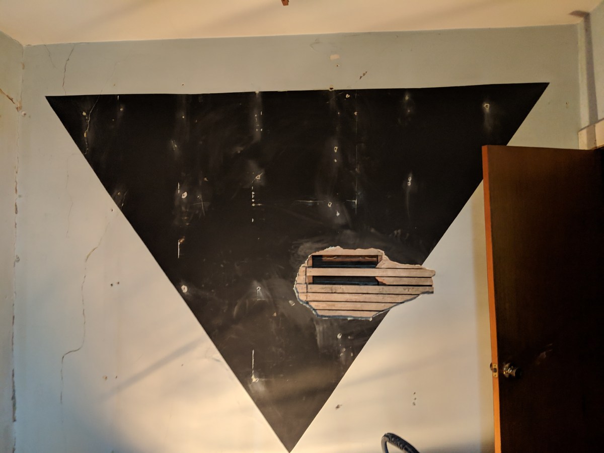 a photo of a black upside down triangle painted on a pale blue wall, taking up most of the wall. it has a lot of holes and patches