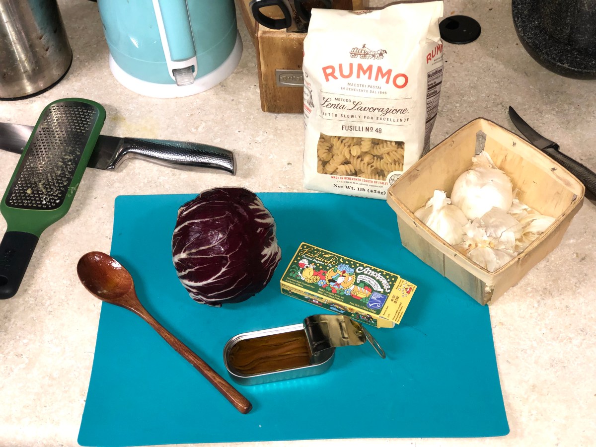 a countertop with a cutting board, a wood spoon, a cheese grater, a Fishwife anchovy box, anchovies in their tin, a bag of uncooked fusili pasta, and a crate of garlic
