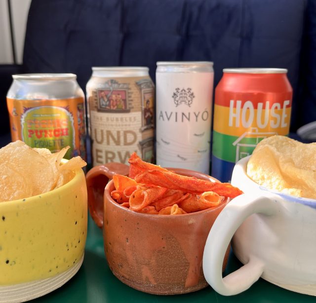 Four canned sparkling wines, and three ceramic mugs filled with different kinds of chips (potato, takis, and salt and vinegar), set against a backdrop of a blue velvet couch.