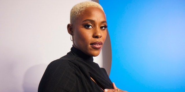 Jerrie Johnson is a Black actor with short cropped blonde hair, they are looking to the camera from a side profile in front of a white and blue background. They have on a black turtleneck