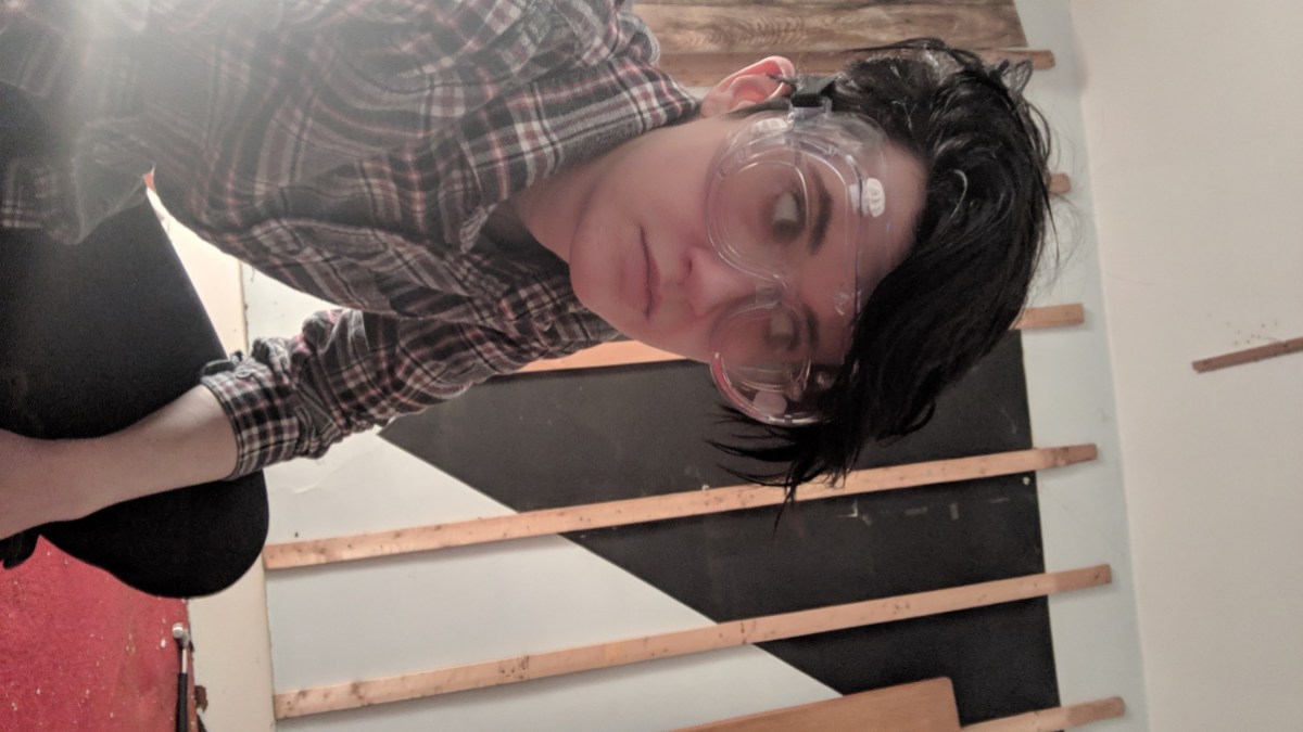 a photo of nico in a flannel and googles crouching in the foreground and giving side eye to a weird black triangle painted upside down and dominating a wall in the background