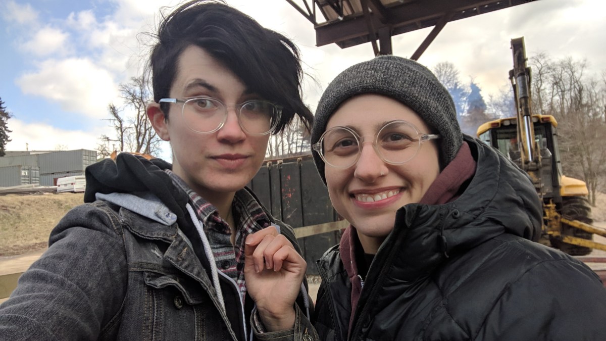 Nico and Sadie smile for a selfie while at the dump. they're both wearing glasses and jackets