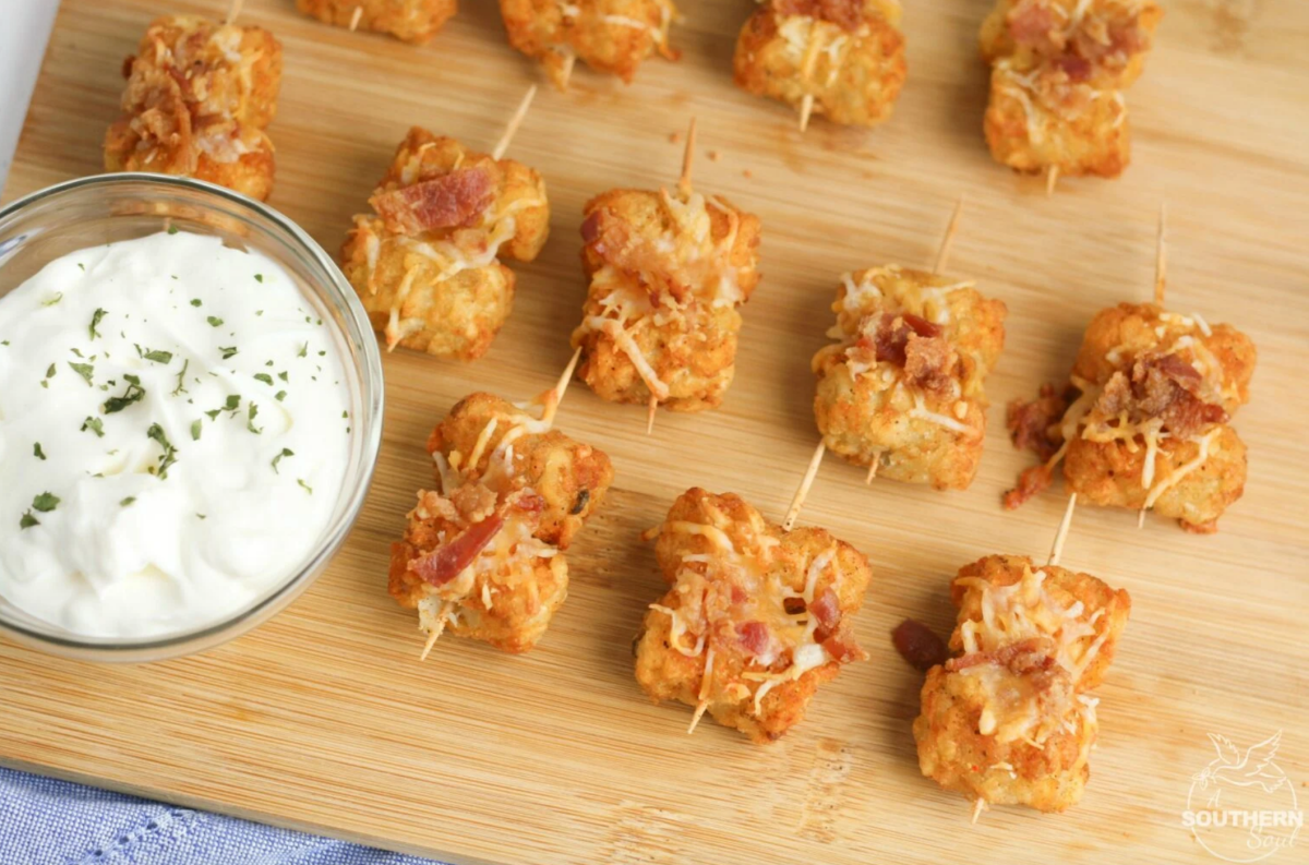 a platter of tater tots covered in toppings on a wood cutting board, next to some white sauce