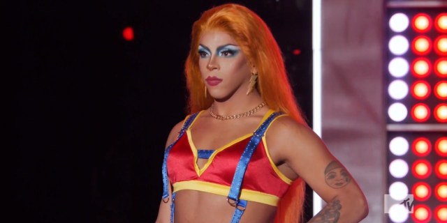 Drag Race 1508 recap: Jax stands on stage in a red and yellow top with blue suspenders and an orange wig.