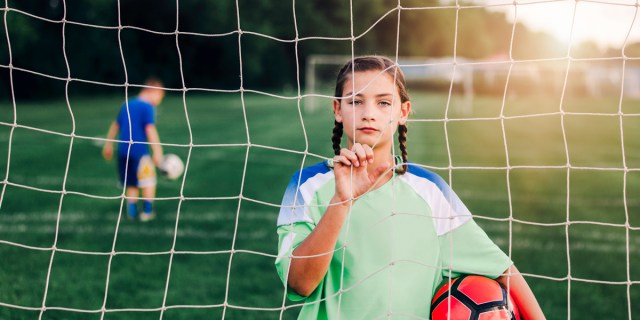 11 year old girl holding soccer ball looking through soccer goal net