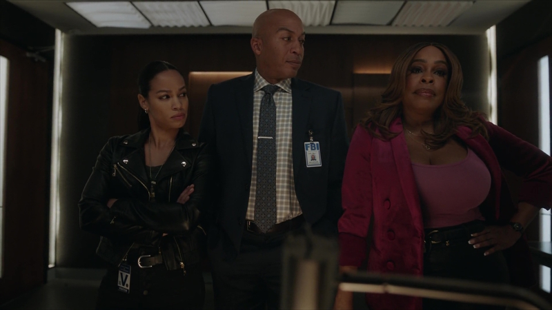 The Rookie: Feds - Naomi and Carter stare in disbelief at Simone while listening to the interrogation fo the murder suspect. Simone is completely unbothered. Naomi is wearing a black leather motorcycle jacket and black jeans, with her visitor's pass affixed to her belt. Carter is wearing a blue suit with grey, brown, and white checkered shirt and tie. His FBI badge is featured prominently on his left breast pocket. Simone is wearing plum double breasted blazer with a lighter plum tank underneath. She has her left hand perched on her hip.
