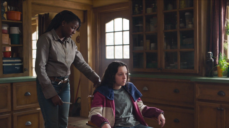 Queer actors Rutina Wesley and Bella Ramsey as Maria and Ellie in The Last of Us: Maria is cutting a grumpy Ellie's hair 