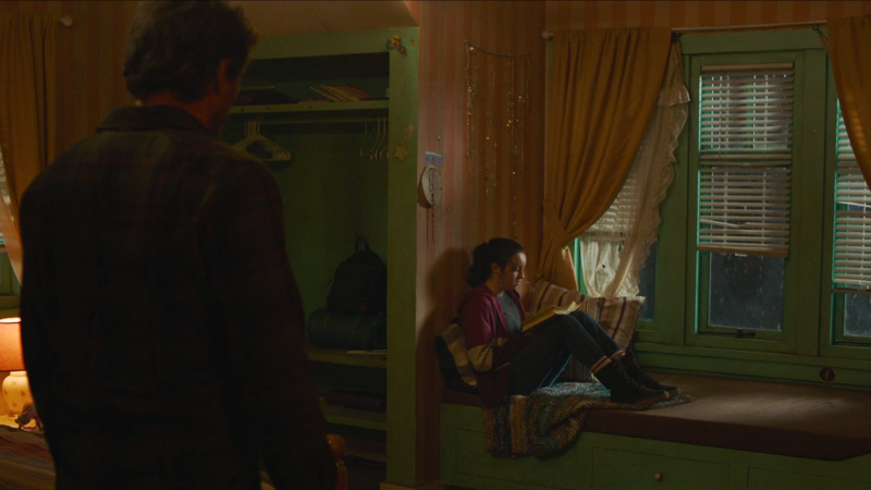 The Last of Us: We see the back of Joel as he stands to the side of the frame, looking at Ellie who is sitting on a window seat reading a teen girl's diary.
