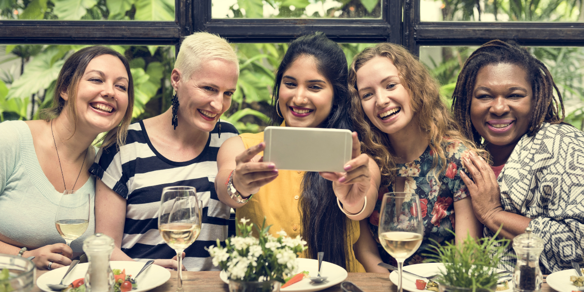 five femmes are hanging out at a gorgeously set table, smiling and taking a group selfie