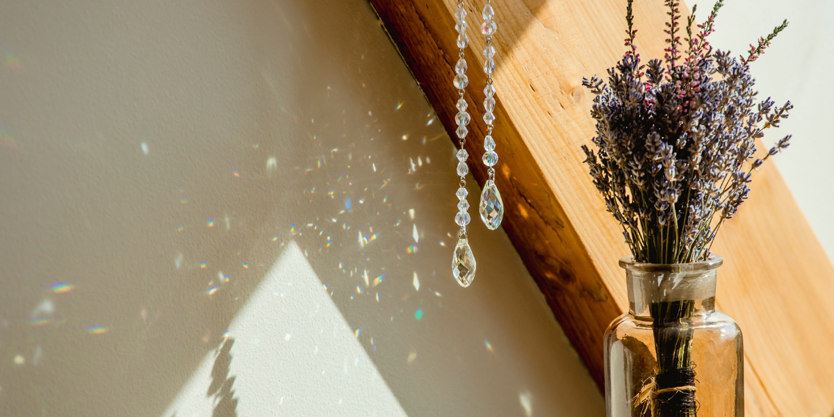 On the right, there is a glass vase filled with lavender and some dried pink flowers. A white wall and a diagonal wood panel are in the background. To the left of the vase, there are two, hanging strings of crystals. They reflect rainbow flecks of light onto the wall.