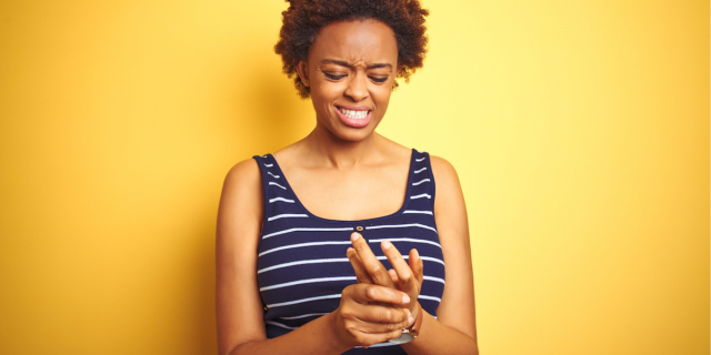 A Black woman with short, curly hair wears a navy blue tank top with white stripes and stands against a yellow background. She holds one hand in the other and grimaces, suggesting that she has chronic pain in her fingers.